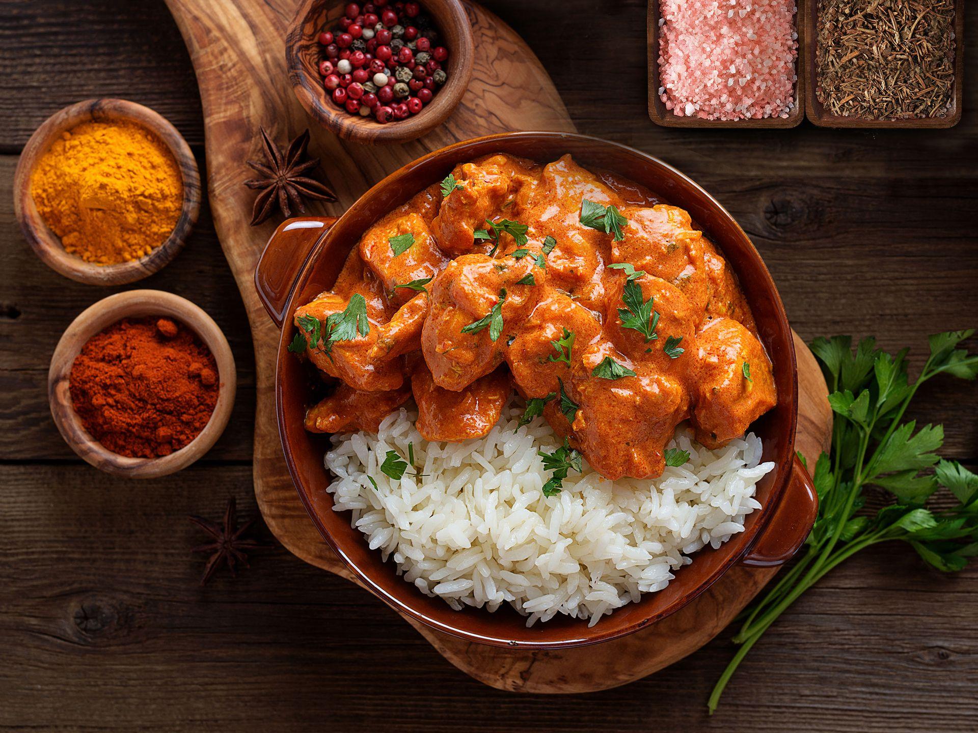 Bowl of chicken curry with rice, surrounded by spices and herbs on a wooden board.